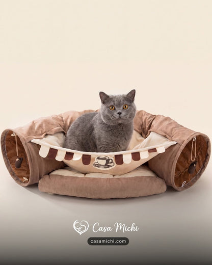 Grey British Shorthair sitting in brown cafe-style cat tunnel bed with coffee cup emblem